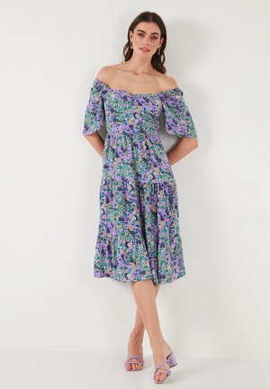 Vestido lavanda de hombros descubiertos con mangas cortas, patrones florales en verde y amarillo, dobladillo con volantes en capas y tejido ligero.