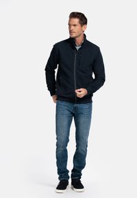 Marine blauwe ritsjack met een gestructureerde stof, hoge kraag en zijzakken, gecombineerd met blauwe jeans en zwarte instapschoenen.
