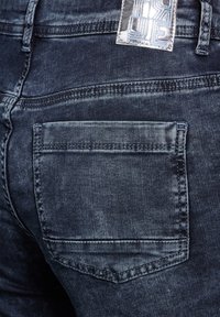 Jeans in denim blu scuro con un design aderente. Presentano una tasca posteriore, leggeri segni di usura e un'etichetta metallica del marchio all'altezza del cinturino.