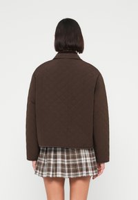 Άτομο με κοντά σκούρα μαλλιά που φοράει σκούρο καφέ καπιτονέ μπουφάν και πλισέ καρό φούστα, γυρισμένο πλάτη σε απλό φόντο.