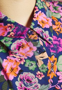 Chemise à fleurs en tissu bleu foncé avec des fleurs vibrantes de couleur rose, orange et verte. Détails notables : un col pointu et des boutons bleus.