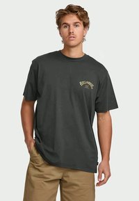 Young man with wavy hair wears dark gray Billabong T-shirt and tan shorts, standing with one hand in pocket against plain background.