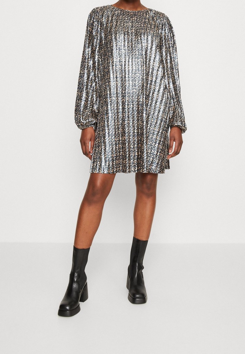 Robe argentée et noire à sequins, coupe ample, manches longues bouffantes et encolure arrondie, associée à des bottines noires à talons.