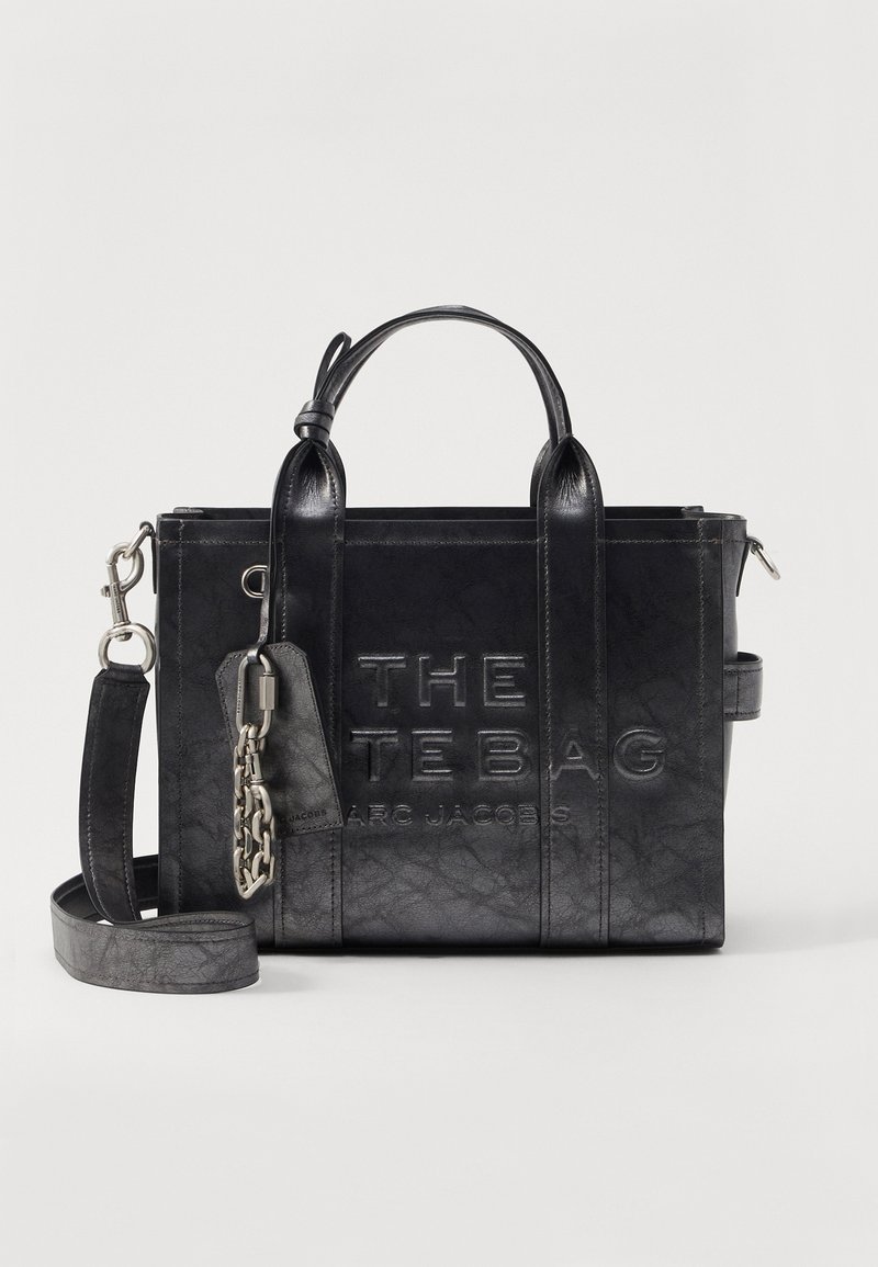 Zwarte leren handtas met twee handvatten, afneembare schouderriem, gegraveerde tekst "THE TEBAG MARC JACOBS" en een metalen ketting sleutelring.
