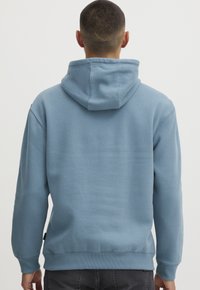 Felpa leggera blu con cappuccio realizzata in tessuto morbido, dotata di un cappuccio ampio, polsini e orlo a coste. Cuciture visibili e una piccola etichetta sul lato.