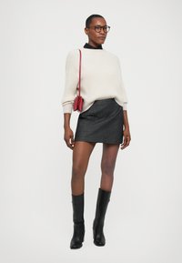 Gebreid crèmekleurig sweater, aansluitende zwarte minirok, zwart kniehoog schoenen, rode schoudertas, bril, staand tegen een effen achtergrond.
