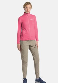 Veste zippée rose vif avec un col haut et une surface texturée, associée à un pantalon beige clair et des chaussures de randonnée bleu marine.