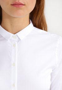 Camisa blanca de botones con cuello puntiagudo, que presenta una textura suave y seis botones visibles en la parte delantera. Diseño simple y clásico.