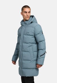 Mėlynas pūkinis paltas su gobtuvu, turintis užtraukiamą kišenę, siuvinėtą tekstūrą ir laisvą prigludimą, pagamintas iš vandeniui atsparios medžiagos.