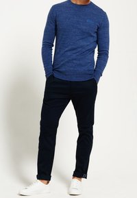 Blå långärmad skjorta med rund halsringning och en diskret logotyp, kombinerad med marinblå byxor och vita sneakers. Avslappnad, figurnära design.