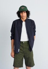 Marineblå skjorte med knapper over en hvit tanktop, parret med olivengrønne shortser. Modellen har på seg en mørkegrønn caps med brodert tekst.