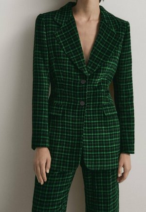 Person wearing a dark green and black plaid blazer and matching pants, standing against a plain light background.