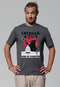 Graues Baumwoll-T-Shirt mit einem grafischen Design, das eine schwarze Katze, einen roten Kreis, das Kapitol und den Text "CATZILLA KÖNIG DER PAWSTER" zeigt. Kurze Ärmel.