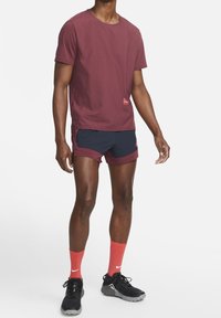 Camiseta atlética granate y pantalones cortos azul marino, con cuello redondo y logotipos. Zapatillas negras con suelas grises. Medias rojas brillantes completan el conjunto.