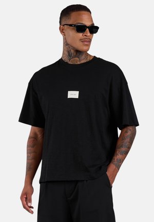 Mann mit Tattoos und Sonnenbrille, der ein schwarzes oversized T-Shirt mit kleinem weißem Logo auf der Brust trägt, posiert mit einer Hand in der Tasche.