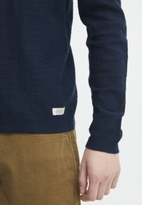 Mornarska majica s dugim rukavima napravljena od mekanog materijala s teksturiranim završetkom; ima malu oznaku brenda na rubu, u kombinaciji s smešnim hlačama.