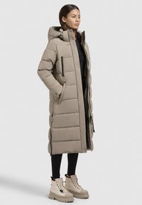 Beige lang dunfrakke med hætte, der har quiltet design, sidelommer og to forlommer. Båret over en sort top og leggings, parret med beige støvler.