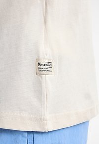 Camiseta de algodón color crema con un dobladillo recto y una etiqueta cosida que dice "Petrolind", ubicada en la parte inferior izquierda.