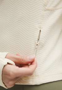 Bluză crem deschis cu fermoar, cu o textură în zigzag, având un fermoar vizibil și un tab de tragere. Detalii cusute și găuri pentru degete pe mâneci sunt prezente.