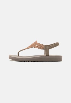 Beige flad sandal med perlebesat overdel, tåstrop og elastisk bagrem på en struktureret sål mod en ensfarvet baggrund.