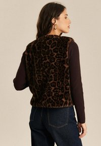 Kobieta w brązowej kamizelce z futra syntetycznego w w „leopardzie” na ciemnym, długim rękawie topie i ciemnoniebieskich dżinsach, stojąca bokiem, aby pokazać tył kamizelki.
