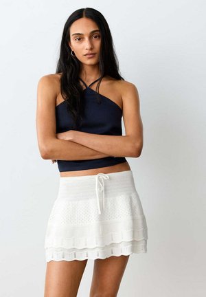 Femme aux longs cheveux foncés portant un top dos nu noir et une jupe blanche tricotée, debout les bras croisés devant un fond uni.