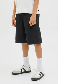 Shorts cargo noirs dotés de multiples poches, fabriqués en tissu coton, avec une coupe décontractée. Associés à des chaussettes blanches et des baskets noir et blanc.