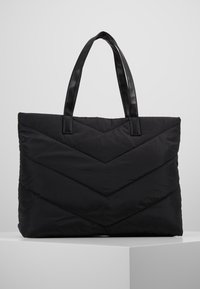 Sac en toile matelassé noir avec couture en chevron et deux poignées en cuir lisse, exposé sur un piédestal blanc contre un fond gris uni.