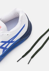 Detailní záběr bílé a modré sportovní obuvi s černými a zelenými šňůrkami umístěnými vedle na bílém pozadí.