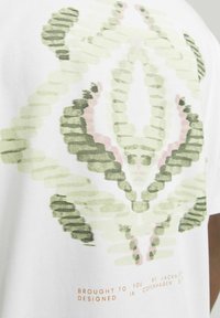Weißes T-Shirt mit einem grünen und rosa abstrakten Design. Der Stoff ist weich, mit bedrucktem Text in Hellbraun am unteren Rand.