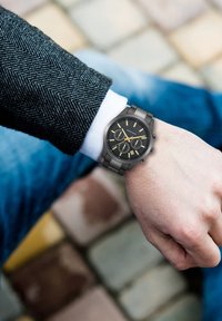 Zwarte roestvrijstalen horloge met een ronde wijzerplaat, gouden accenten, drie chronograaf sub-dialen en een datumscherm, gedragen aan een pols in de textuur van een pakmouw.