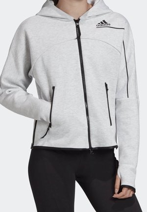 Jasnoszara bluza z kapturem i zamkiem błyskawicznym z czarnymi suwakami, logo Adidas na piersi oraz czarne spodnie, noszona przez osobę z jedną ręką w kieszeni.