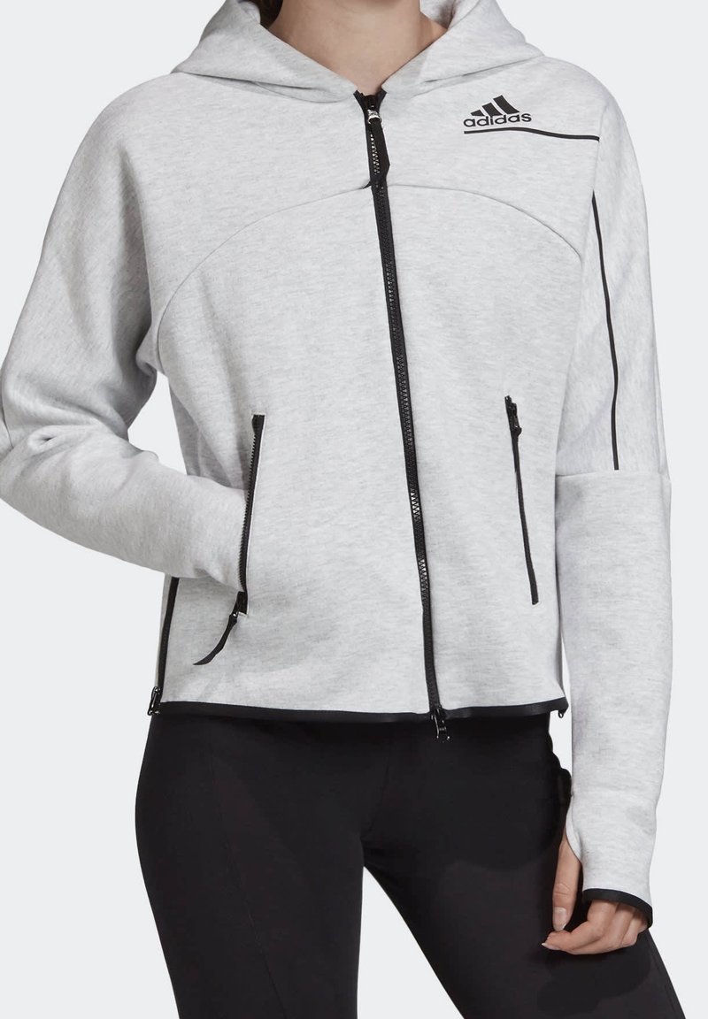 Veste à capuche gris clair zippée avec des fermetures éclair noires, logo Adidas sur la poitrine, et pantalon noir, portée par une personne avec une main dans la poche.