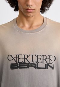 Gray, short-sleeve T-shirt made of soft fabric, featuring a large black graphic design with "VERTERE" and "BERLIN" text at the center.