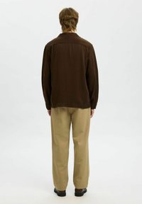 Brązowa koszula z długim rękawem i spicznym kołnierzykiem, w luźnym kroju, z zapięciem na guziki przy mankietach, zestawiona z jasnobeżowymi spodniami i czarnymi butami.