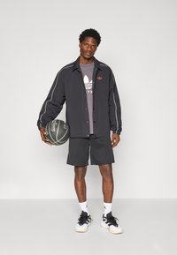 Veste de sport noire avec des accents blancs, t-shirt gris arborant un logo, shorts noirs et baskets noir et blanc, tenant un basketball.