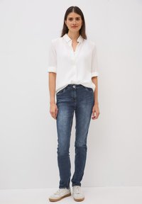 Chemise blanche à manches retroussées, associée à un jean skinny bleu foncé et des baskets blanches avec des semelles beiges, se tenant contre un fond uni.