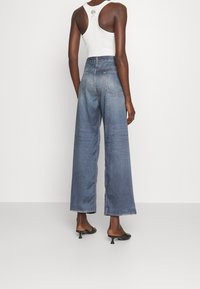 Jean en denim taille haute et jambes larges, bleu moyen, avec poches arrière classiques et ourlet brut. Associé à un haut sans manches blanc.