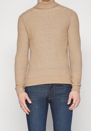 Pull à col roulé côtelé beige en maille texturée, avec des manches longues et une coupe ajustée. Porté avec un jean bleu foncé.