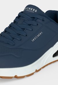 Zbliżenie na granatowy sneaker Skechers ze sznurowadłami, białą podeszwą pośrednią i brązową podeszwą zewnętrzną, z widoczną nazwą marki na języku i boku.