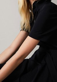 Sort kjole med krave, korte ærmer, glat stof, indsnævret talje med en bundet accent. Minimalistisk design med en ren silhuet.