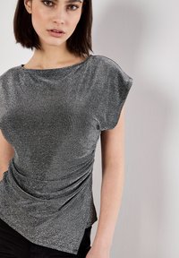Silberner metallischer Top mit kurzen Ärmeln, drapierter Stoff, der einen asymmetrischen Saum bildet, glatte Textur und leichter Glanz vor einem neutralen Hintergrund.