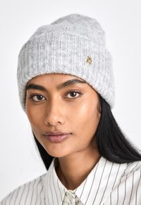 Harmaa neulepipo, jossa on ribbikuvio ja käännetty resori, jossa on pieni kultainen logotehoste. Yhdistetty raitapaidan kanssa.