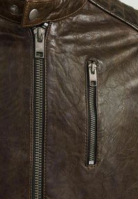 Nahaufnahme einer braunen Lederjacke mit einer Reißverschlusstasche und einem metallischen Reißverschluss. Abgenutzte Textur mit Nähdetails entlang der Kanten.