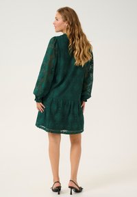 Grön spetsklänning med långa, puffiga ärmar, blommönster och lager på lager-skirt. Har en rund nacke och en svävande, lätt textur.