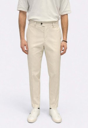 Uomo che indossa pantaloni beige su misura, sneakers bianche e una camicia bianca a maniche corte, in piedi davanti a uno sfondo bianco uniforme.