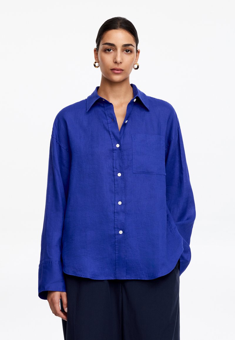 Femme aux cheveux foncés attachés, portant de grandes boucles d'oreilles en or et une chemise bleue ample à boutons avec une poche poitrine.