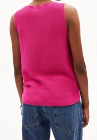 Person klädd i en ärmlös ljusrosa texturerad topp och blå denimjeans, sedd bakifrån mot en vit bakgrund.