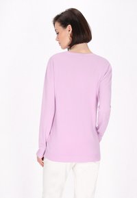 Maglione lilla con maniche lunghe e vestibilità rilassata. Texture liscia e orlo a coste. Il modello è in piedi con la schiena rivolta, indossando pantaloni bianchi.