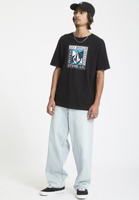Sort kortærmet t-shirt med et grafisk logo i blå og hvid. Kombineret med lyseblå baggy jeans og sorte sneakers. Bærer en sort kasket.